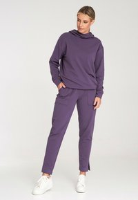Fioletowa bluza z kapturem i pasujące spodnie z miękkiego materiału, z bocznymi kieszeniami i rozcięciami przy kostkach, w zestawie z białymi trampkami.