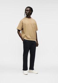 Μπεζ βαμβακερό t-shirt με τσέπη, συνδυασμένο με σκούρο μπλε παντελόνι με κοψίματα και λευκά αθλητικά παπούτσια. Απλός σχεδιασμός και άνετη εφαρμογή.