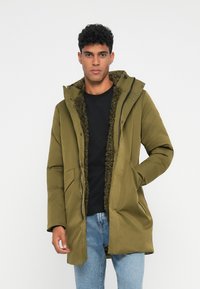 Junger Mann in einer olivgrünen Kapuzenjacke mit Fleecefutter, schwarzem Shirt und blauen Jeans, der vor einem schlichten Hintergrund steht.