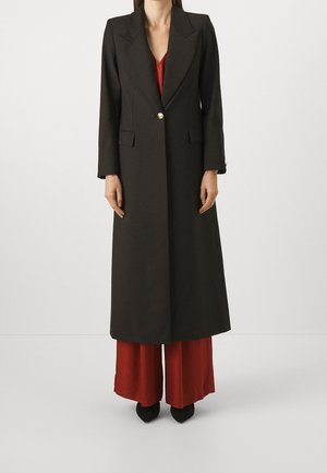 Femme portant un long manteau noir à un bouton unique sur un pantalon rouge à jambes larges et des chaussures noires pointues, debout sur un fond blanc uni.