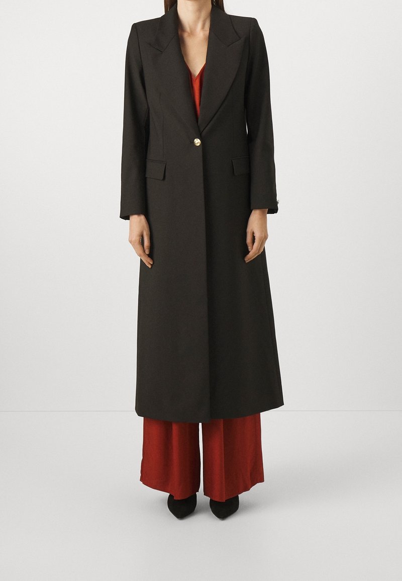 Femme portant un long manteau noir à un bouton unique sur un pantalon rouge à jambes larges et des chaussures noires pointues, debout sur un fond blanc uni.