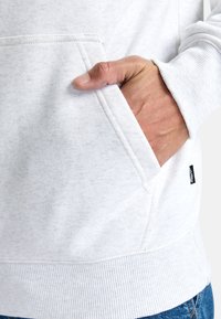 White hoodie with a soft, textured finish. Features a front kangaroo pocket and ribbed cuffs. Small black brand label on the side seam.