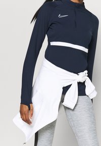 Tmavě modrý sportovní top s dlouhým rukávem, zipovým stahovacím límcem a bílým horizontálním pruhem. Bílá mikina uvázaná v pase, doplněná šedými legínami.