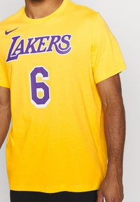 Żółta koszulka z bawełny, z fioletowym napisem "LAKERS" i dużą liczbą "6" w kolorze fioletowym z białymi akcentami. Krótkie rękawy, okrągły dekolt.