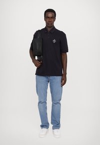 Marineblå polo skjorte med en liten hvit logo, parret med lyseblå jeans og hvite sneakers. Svart crossover-veske slengt over skulderen.