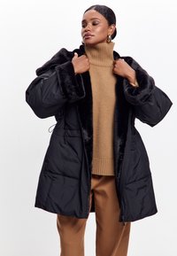 Sort dunjakke med et faux fur-krave, med lynlås lukning, to forlommer og knælang pasform, lagd over en beige sweater.
