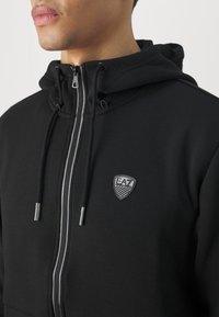 Felpa nera con zip, realizzata in un tessuto texturizzato, dettagli grigi sulla zip e logo EA7 ricamato sul petto sinistro. Coulisse regolabile per il cappuccio.