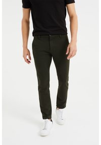 Dark green patterned trousers made of textured fabric, tailored fit, with a zip fly, side pockets, and worn with white sneakers.