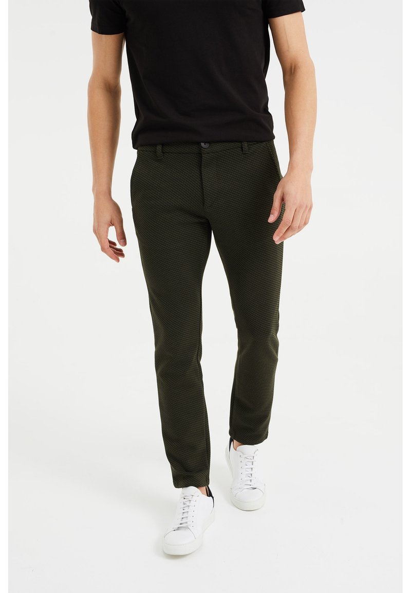 Dark green patterned trousers made of textured fabric, tailored fit, with a zip fly, side pockets, and worn with white sneakers.