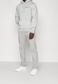 Jasnoszara bluza z kapturem z przednią kieszenią i sznurkiem ściągającym, połączona z dopasowanymi szarymi dresami i białymi sneakersami. Posiada haftowane logo.