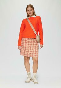 Pull en maille matelassée orange avec des manches longues, chemise blanche à col en dessous, jupe à motif pied-de-poule, sac bandoulière beige et bottes crème.
