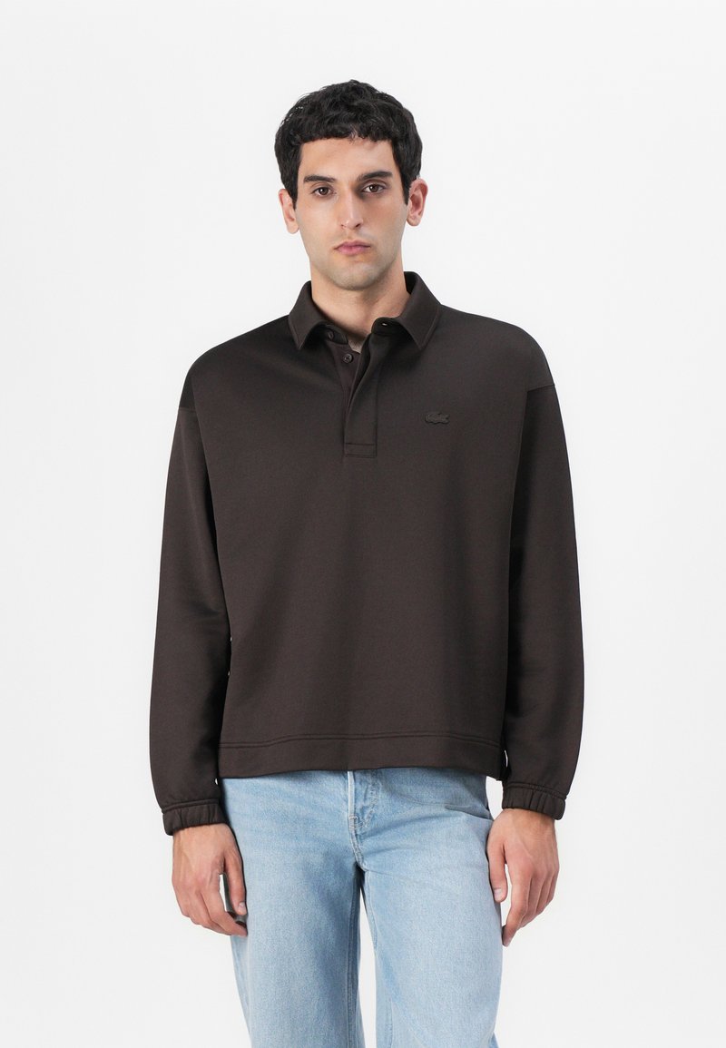 Felpa polo marrone con colletto e polsini a costine, dotata di un piccolo logo sul petto e una vestibilità rilassata. Abbinata a jeans in denim chiaro.