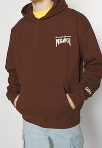 Hnědá bavlněná mikina má přední klokaní kapsu, bílé logo "PEGADOR" na hrudi a detail "500" na rukávu; má volný střih a kapuci.