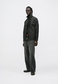 Black lightweight jacket with two chest pockets, paired with dark gray loose-fitting jeans and black shoes, worn by a model.