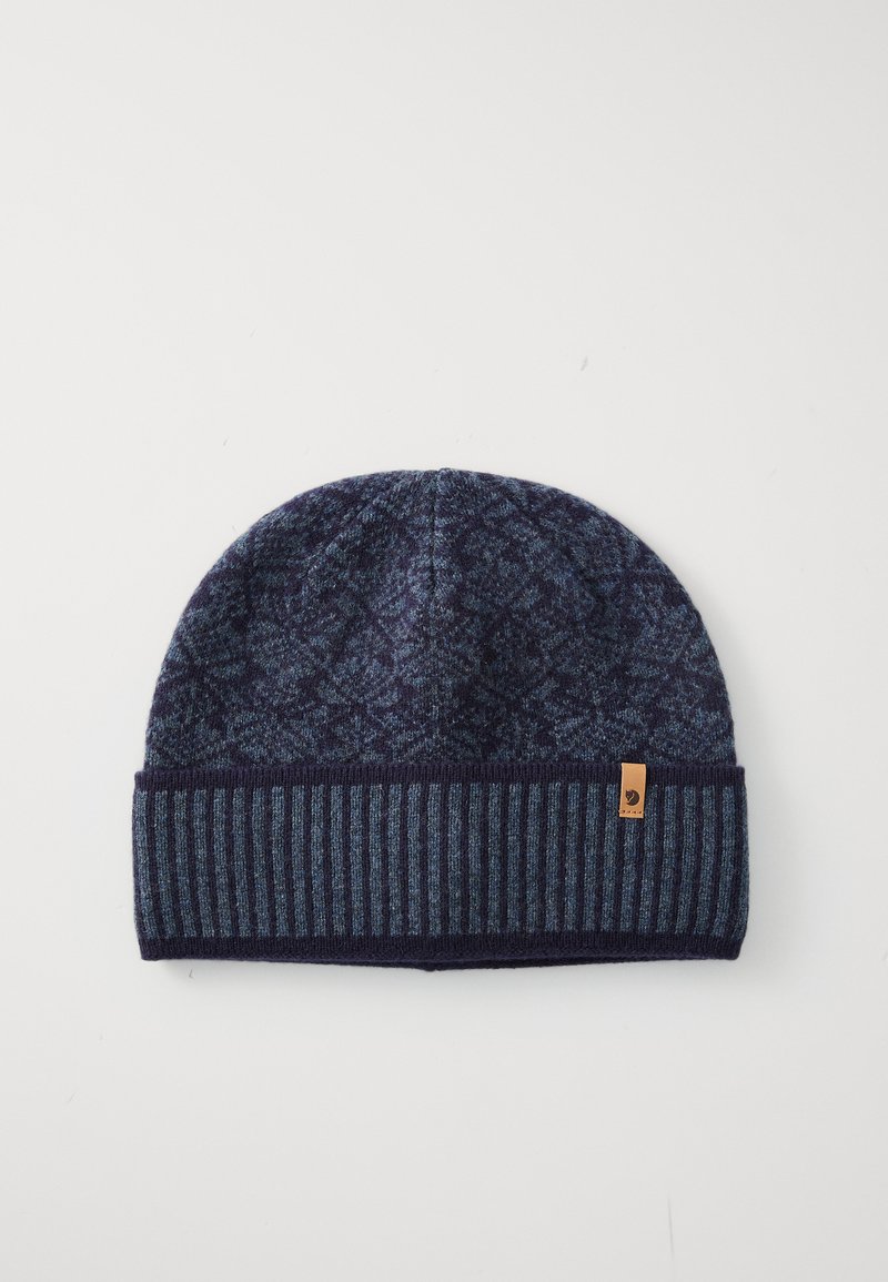 Tumed sinine kudum beanie mustriga peal ja ribakantud äärega, millel on väike pruun logo silt valgel taustal.