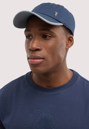 Joven con gorra azul marino con un pequeño logo de pájaro y camiseta a juego azul marino con un diseño abstracto de un perro.