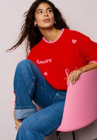 Rode gebreide trui met roze accenten, met de opdruk "Amore" en hartmotieven. Spijkerbroek met omgeslagen zoom. Zit op een roze stoel.