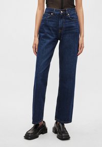 Tumši zili džinsu džinsi ar taisnu kāju griezumu, augstu vidukli, priekšējām kabatām, vara pogas aizdari un kontrastējošām šuvju detaļām.