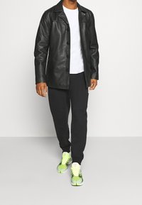 Schwarze Lederjacke mit Knopfverschluss, über einem weißen T-Shirt. Kombiniert mit schwarzen Hosen und neongrünen Sportschuhen.