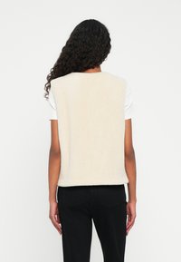 Cremiger Fleece-Weste mit kastiger Form, abgerundeten Schultern und weicher Textur, getragen über einem weißen Hemd und kombiniert mit schwarzen Hosen.