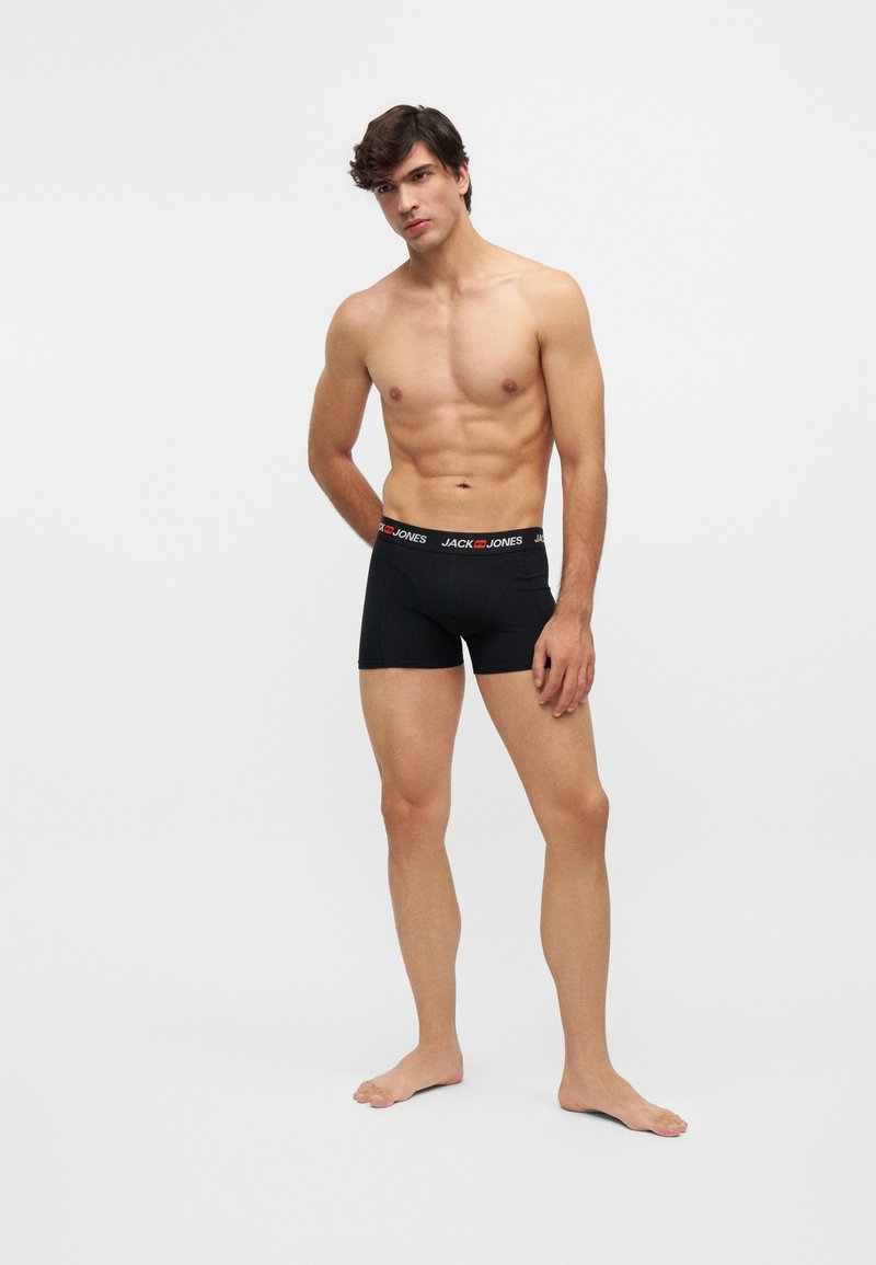 Hombre sin camiseta, de pie descalzo, con bóxers negros Jack & Jones sobre un fondo liso y claro.