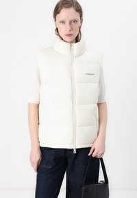 Witte gewatteerde vest met een hoge kraag, ritssluiting en logo-detail. Gedragen over een T-shirt met korte mouwen, gecombineerd met donkere denim en een zwarte tas.