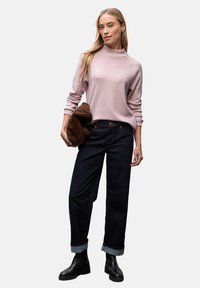 Pull en tricot rose clair avec un col montant, assorti à un jean bleu foncé à manches retroussées. Le modèle tient un sac en daim marron. Bottines noires.