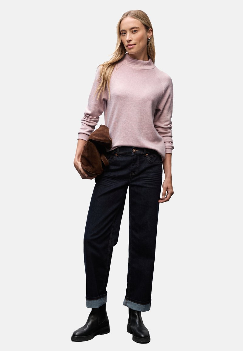 Pull en tricot rose clair avec un col montant, assorti à un jean bleu foncé à manches retroussées. Le modèle tient un sac en daim marron. Bottines noires.