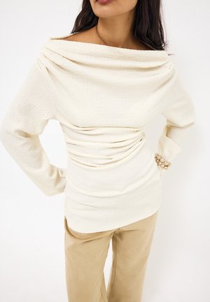 Kobieta ubrana w kremową, teksturowaną bluzkę z długim rękawem i dekoltem w drapowaniu oraz beżowe spodnie, stojąca z rękami na biodrach na białym tle.
