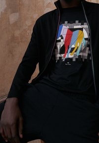 Svart zip-up jacka över en svart t-shirt med en färgglad abstrakt hunddesign på en rutad bakgrund. Mjuk tyg, avslappnad passform.