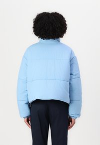 Osoba s kudrnatými vlasy, která má na sobě světle modrou péřovou bundu a tmavé kalhoty, stojící čelem k jednobarevnému bílému pozadí.