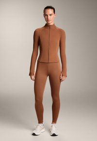 Top aderente marrone con zip e maniche lunghe, abbinato a leggings coordinati realizzati in materiale liscio e elastico; indossato con scarpe da ginnastica bianche.