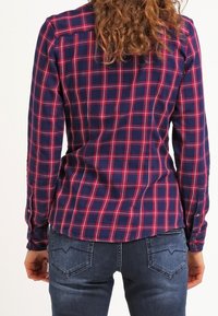 Chemise à carreaux bleu marine et bordeaux avec des manches longues, un ourlet arrondi et une coupe ajustée. Le tissu semble lisse avec une légère brillance.