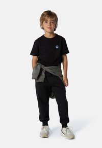 Camiseta negra con un logo azul, sudadera gris atada a la cintura, pantalones jogger negros y zapatillas deportivas beige.