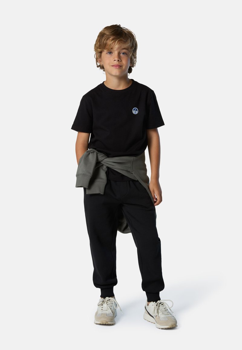 Camiseta negra con un logo azul, sudadera gris atada a la cintura, pantalones jogger negros y zapatillas deportivas beige.