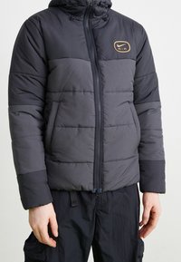 Padded jacket w kolorze ciemnoszarym z zamkiem z przodu i regulowanym kapturem. Ma złote logo Nike Air na klatce piersiowej oraz pikowaną teksturę.
