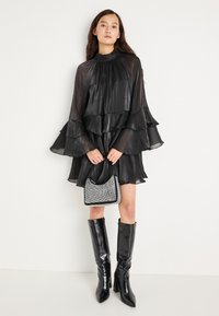 Must mustriga mustriga kleit rippuvate varrukatega, läikiva tekstuuriga. Koos väikeselt metalset käekotiga ja kuni põlveni ulatuvate mustade saapadega.