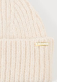 Caciula tricotat�ă de culoare crem, realizată din fir moale, cu textură ribbed vertical; prezintă un mic tag cu logo-ul Barbour de culoare aurie pe manșetă.