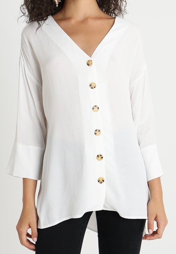 Blouse blanche à manches longues avec col en V, dotée de six boutons bruns sur le devant, portée avec un pantalon noir.