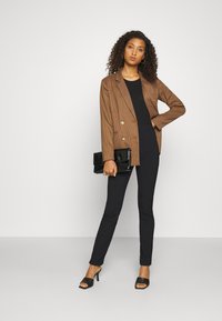 Blazer xadrez castanho sobre uma blusa preta, combinado com calças pretas justas e sandálias pretas de salto. O modelo segura uma clutch preta.