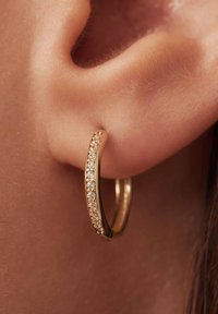 Boucle d'oreille en or avec une surface texturée et des pierres précieuses transparentes incrustées le long du bord avant, fixée à une oreille percée.