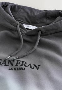 Hanorac gri cu siret, având un logo imprimat "SAN FRAN CALIFORNIA". Materialul are o textură moale și o finisare netedă.
