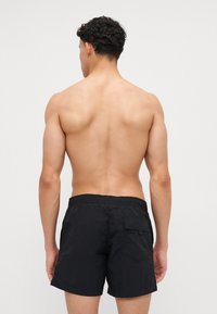 Shorts de bain noirs avec une ceinture élastique à la taille, une poche arrière unique et un tissu léger, présentés de dos sur un torse nu.