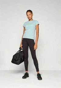 Lyseblå tettsittende t-skjorte med grønt logo, sammen med svarte tights med hvit tekst. Modellen holder en svart treningsbag og har på seg svarte joggesko.