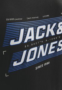 Sort bomulds T-shirt med fed blå og hvid tekstgrafik, der siger "JACK & JONES" med koordinater og årstal nedenfor.