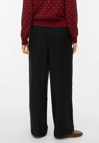 Une personne portant un pantalon noir à jambes larges, un pull bordeaux à pois blancs et des mocassins marron, vue de dos sur un fond blanc.