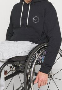 Felpa navy con lacci bianchi e grafica testuale circolare; indossata sopra pantaloni della tuta grigi chiari mentre si è seduti su una sedia a rotelle.