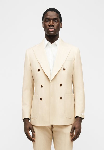 Άνδρας που φοράει ρεβέρ σακάκι διπλής σειράς σε μπεζ χρώμα με παντελόνι που ταιριάζει και λευκό πουκάμισο, μπροστά σε απλό φόντο.