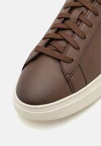 Barna bőr sneaker, kerek orral, kontrasztos fehér gumitalppal és lapos barna fűzőkkel, sima felszínen.