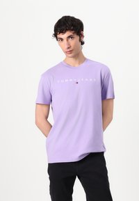 Helllila Baumwoll-T-Shirt mit Rundhalsausschnitt, kurzen Ärmeln und "TOMMY JEANS"-Logo in Weiß mit einem kleinen roten und blauen Emblem.